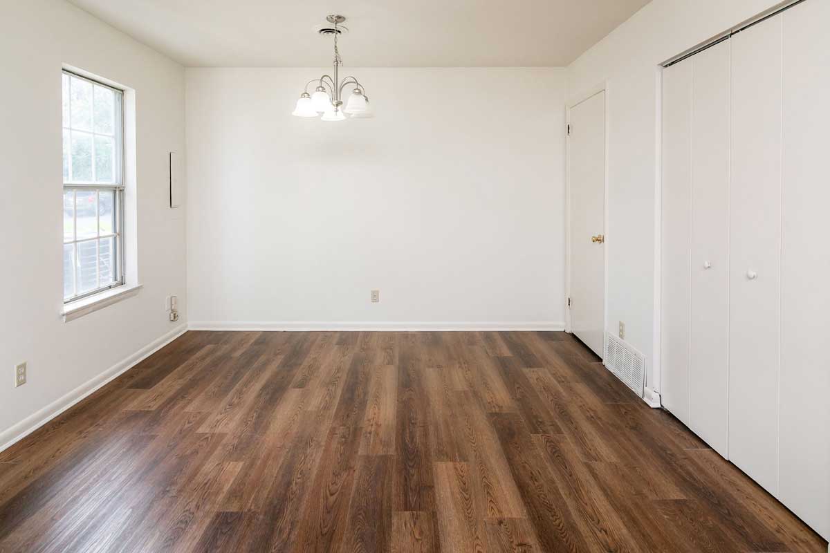 Hickory Hill Townhomes view of eat in area from kitchen. Large empty space with white walls, window on left, utility closet door on right as well as laundry door. A 5 - bulb/globe light fixture hangs in the center of eat in space
