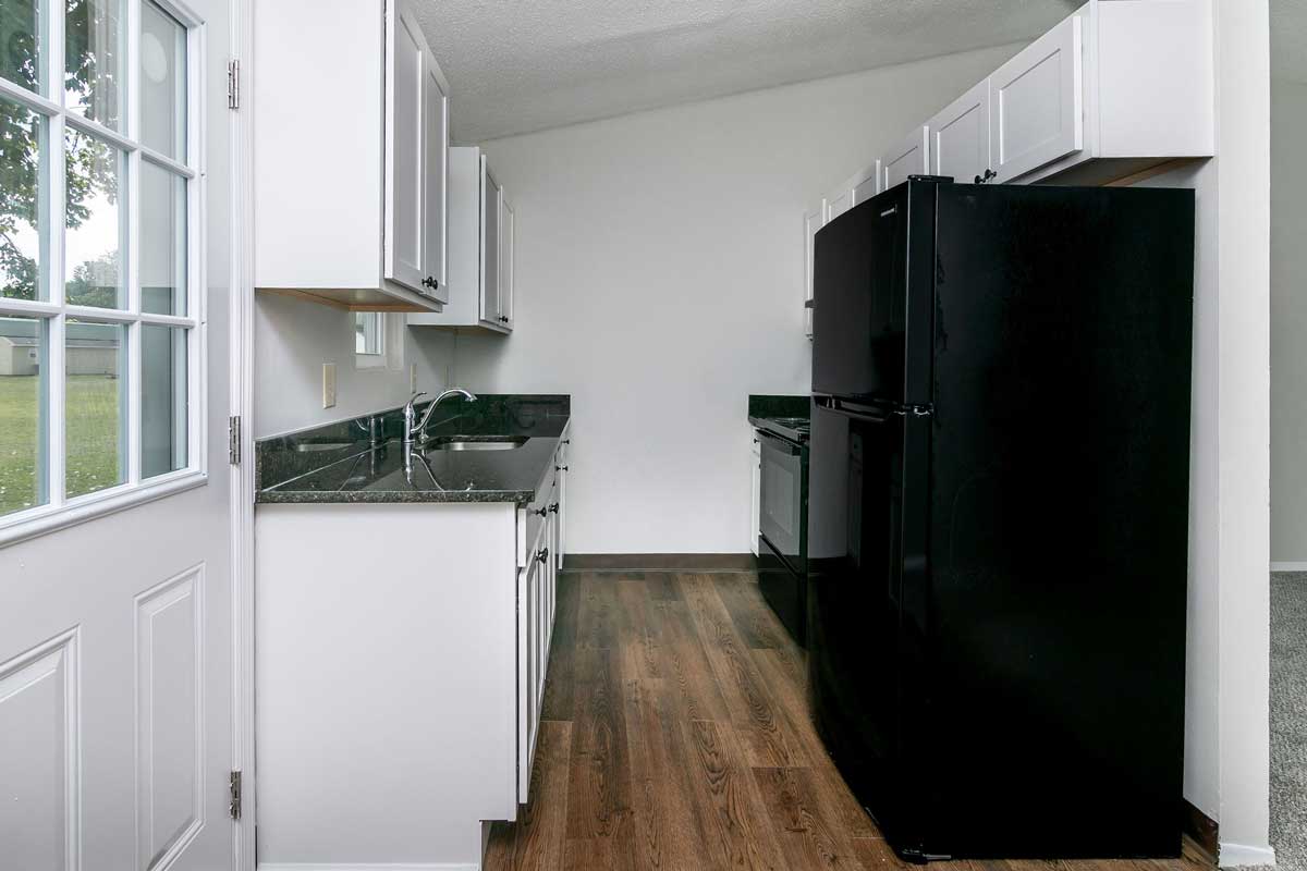 Township kitchen view of entire kitchen looking in. On the left is the back door, cabinets/counter with kitchen sink and window above the sink. Right side, you see black refrigerator, upper white cabinets. stove Hard wood looking vinyl plank floor