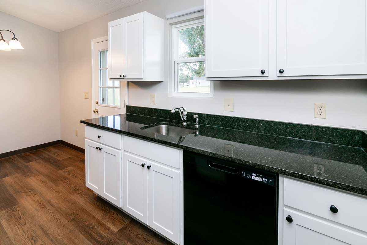 Township kitchen opposite the refrigerator and stove is sink and dishwasher, white cabinets both upper and lower, with black hardware, black granite counter tops and a window above the sink. Door to back yard