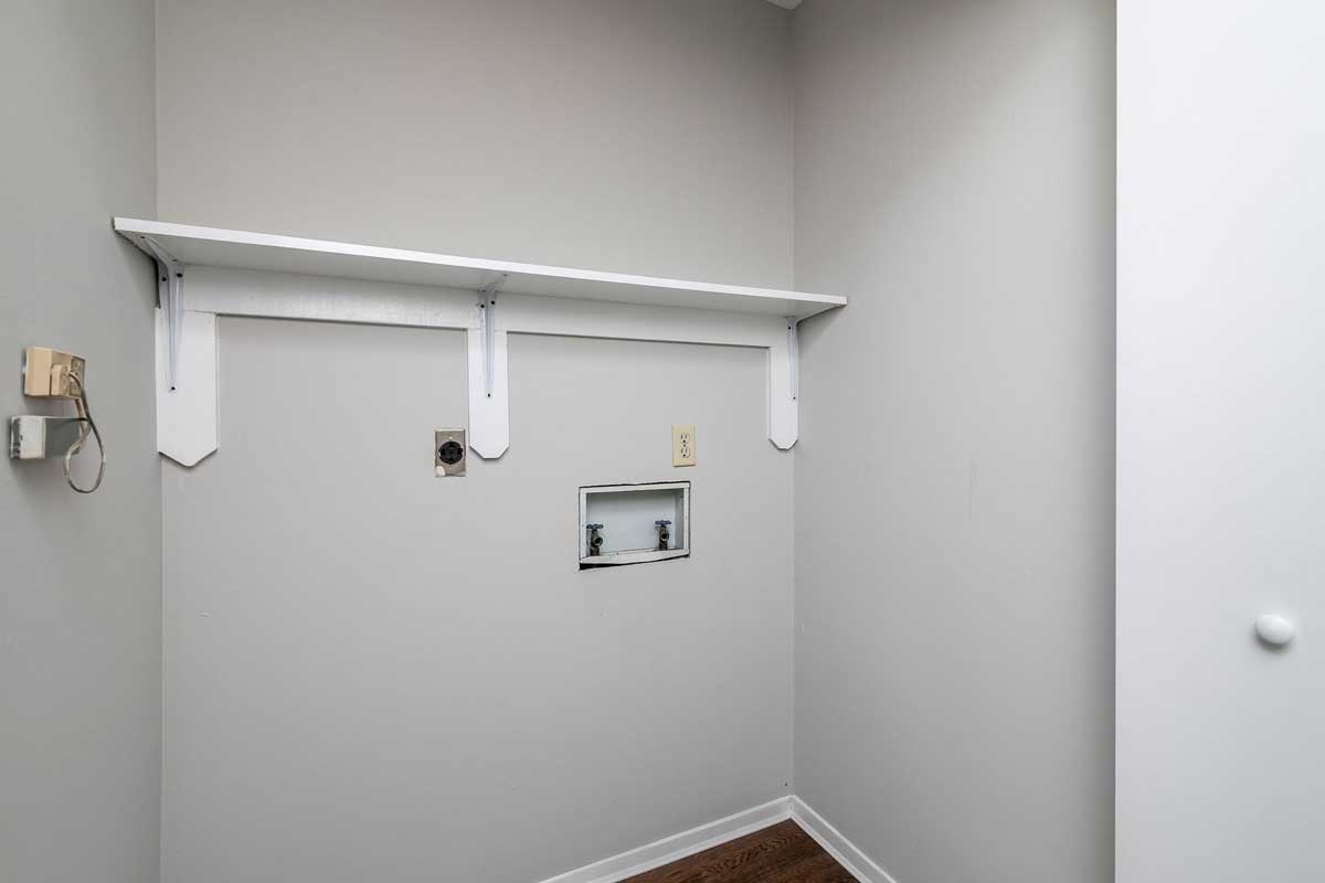 bifold doors located in halllway open to washer and dryer hook up, with white wood shelf installed above for cleaning and laundry supplies